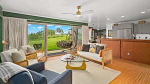 A spacious living room with a large sliding glass door leading to a garden, a sofa, a coffee table, and a kitchen area with cabinets and a countertop.