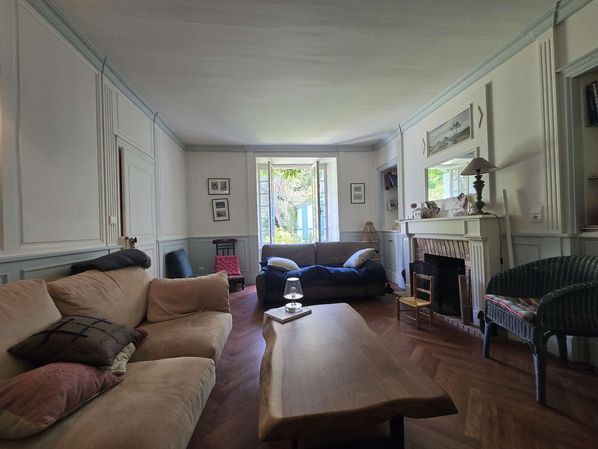 En stue med et træ sofabord, en beige sofa, en blå sofa og en fletstol. Der er en pejs med en lampe på pejsestativet.