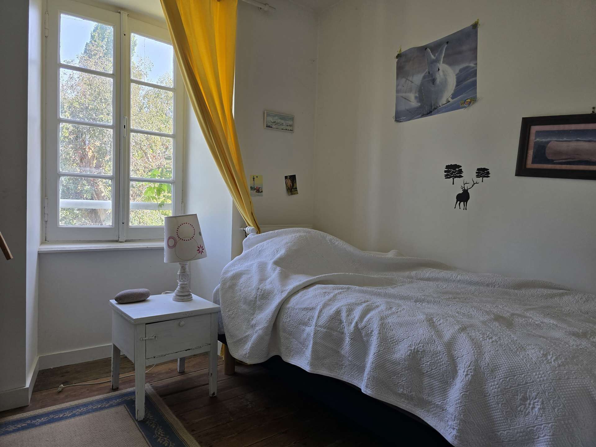 Une chambre à coucher comprenant un lit, une table de chevet avec une lampe, une fenêtre avec des rideaux et des éléments de décoration murale, notamment un tableau et un autocollant représentant un cerf.