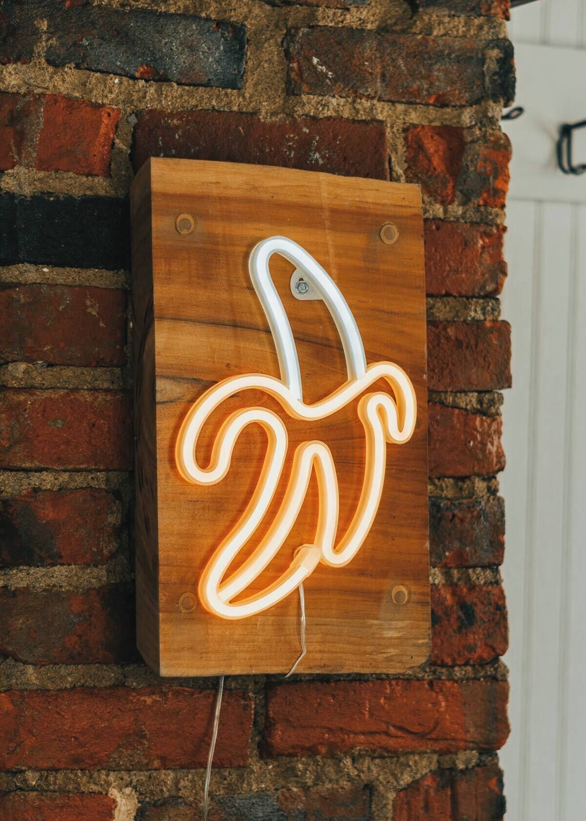 Une enseigne au néon en forme de banane, fixée sur une planche de bois, est accrochée à un mur de briques.