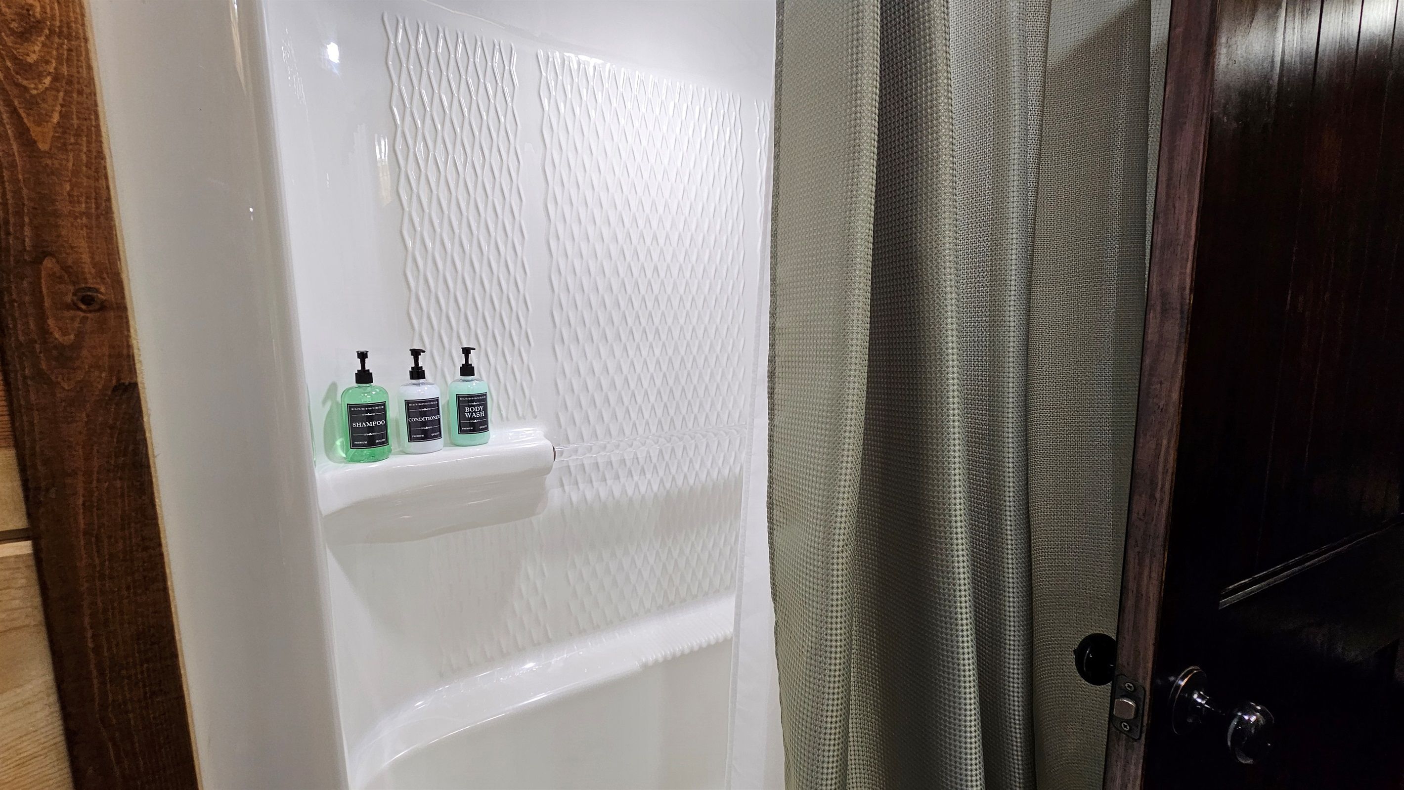A bathroom with a white bathtub, a shelf with three bottles of shampoo, conditioner, and body wash, and a wooden door.