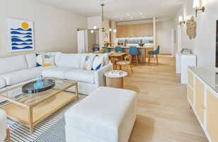 A modern living room with a sofa, coffee table, and ottoman. There is a dining area with chairs and a table. The kitchen is visible in the background with wooden cabinets and a countertop.
