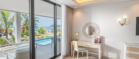 A modern hotel room with a large sliding glass door leading to a pool area, a desk with a chair, a round mirror, and a small cabinet.