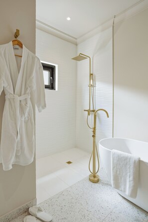 Ein modernes Badezimmer mit einer freistehenden Badewanne, einer goldfarbenen Duschbrause und einem Bademantel, der an einem Holzaufhänger hängt.