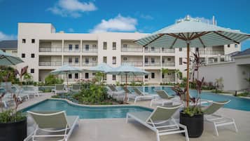 Zona de piscina de hotel con múltiples sillones de descanso y sombrillas, rodeada por un edificio con balcones.
