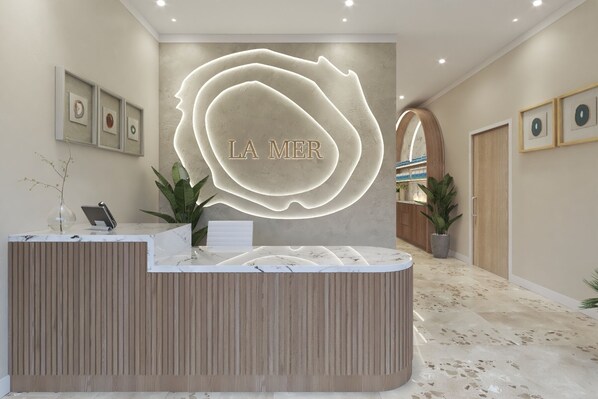 A reception area with a marble counter, a large illuminated 'LA MER' sign, framed artwork on the walls, and potted plants. - Sapphire Sands by Bay Gardens Resorts (Gros Islet)