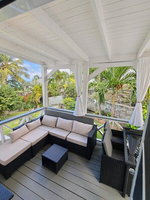 A covered outdoor seating area with a sectional sofa and chairs, overlooking a garden with palm trees and a pool.