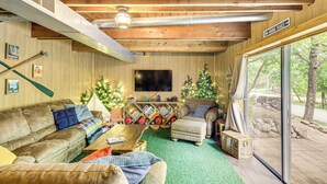 A cozy cabin interior with a wooden ceiling, a flat-screen TV, a fireplace, and a large window overlooking a forested area. - Canal Across Street: Family Cabin in Breezy Point! (Pequot Lakes)