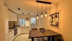 A compact kitchen with modern appliances, a dining area with a wooden table and chairs, and a wall-mounted shelf with decorative items. - Holiday home with air conditioning (Jadranovo)