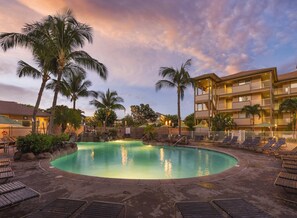 Zona de piscina en un resort con piscina circular, sillas de descanso, palmeras y un edificio de varios pisos con balcones.