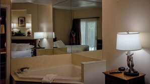 A hotel bathroom with a large mirror, a bathtub, a wooden side table with a lamp, and a remote control.