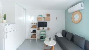 A compact living space with a kitchenette, a flat-screen TV, a small round table, and a grey sofa.