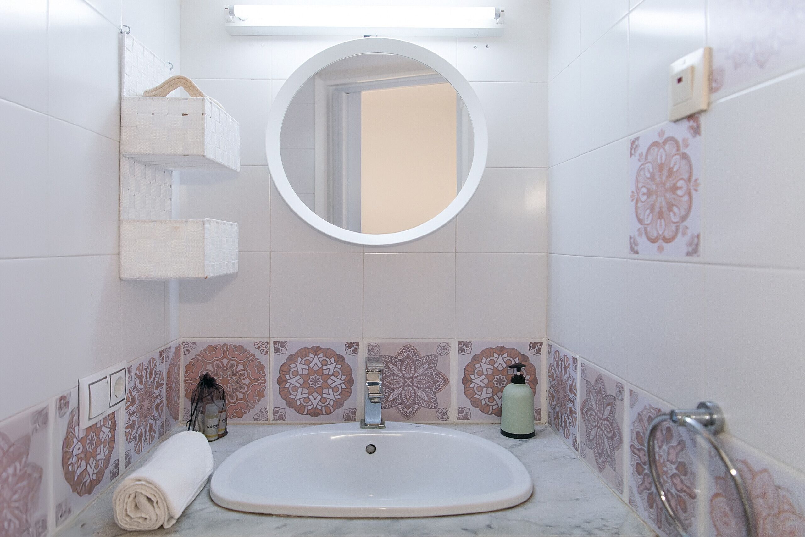 A bathroom with a white sink, a round mirror, patterned tiles, a towel, and a soap dispenser.