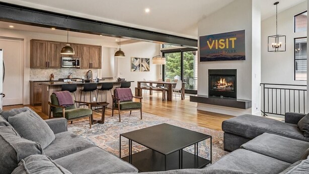 A modern living room with a fireplace, a dining area, and a kitchen visible through an open door. - 120 Cedar Dr | Slopeside, Hot Tub, Mtn Views (Winter Park)