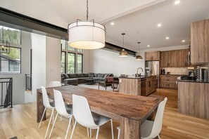 A modern kitchen with a wooden island, white chairs, pendant lighting, and a view of a green landscape.