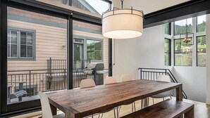 A dining area with a wooden table and chairs, a hanging light fixture, and large windows offering a view of the outdoors. - 120 Cedar Dr | Slopeside, Hot Tub, Mtn Views (Winter Park)