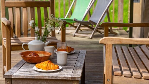Une table en bois sur laquelle sont disposés des aliments pour le petit-déjeuner, notamment des croissants, un bol et une théière. On aperçoit deux chaises en bois et deux fauteuils verts à l’arrière-plan.