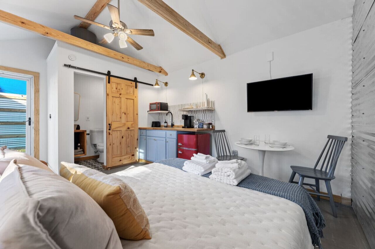 A bedroom with a large bed, a wooden ceiling, a sliding door, a kitchenette, a TV, and a small table with chairs.