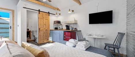 Een slaapkamer met een groot bed, houten plafond, een schuifdeur, een kitchenette, een televisie en een klein tafeltje met stoelen.