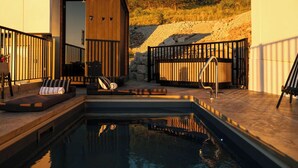 A modern outdoor pool area with a wooden deck, a seating area with cushions, and a view of a forested hillside.