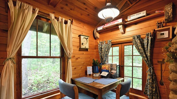 Interior de una cabaña de madera con una mesa de comedor, dos sillas, un reloj y cortinas.