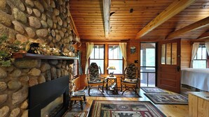 Interior acogedor de una cabaña con pared de piedra, techo de madera, chimenea, sillones mecedores y una alfombra grande.