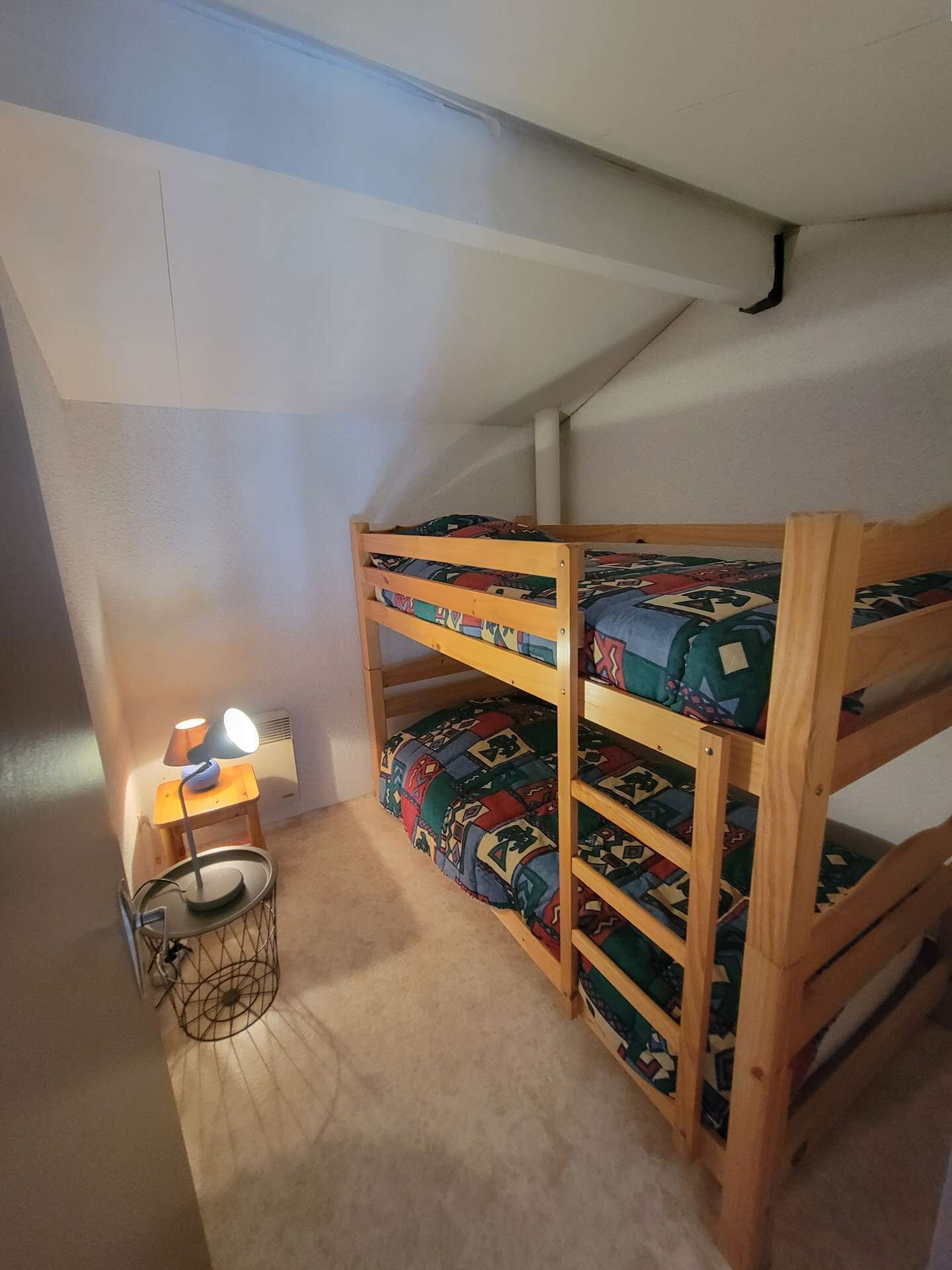 A bunk bed with patterned bedding in a room with a sloped ceiling and a small table with a lamp.