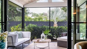 An open-plan living area with a white sofa, a round coffee table, and a dining table. There is a view of a backyard with a wooden fence and greenery.