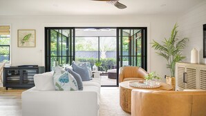 A modern living room with a white sofa, a brown leather chair, and a coffee table. There is a ceiling fan, a large window, and a view of the outdoors.