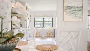 A bright, white interior with a dining area featuring a round table and two chairs, a kitchen area with white appliances, and a large window with a view of greenery.