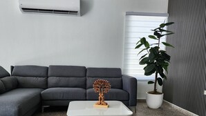 Una sala de estar moderna con un sofá seccional gris, una mesa de centro blanca con base de madera, una escultura de árbol decorativa, una planta en maceta y una pared a rayas.