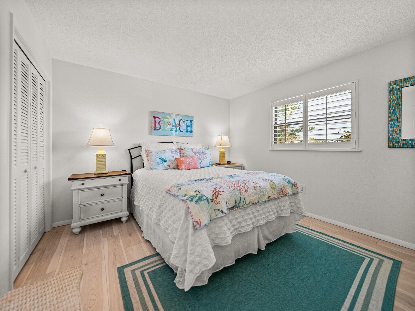 A bedroom with a bed, two bedside tables, a nightstand, a window with blinds, a wall-mounted artwork, and a door with louvered shutters.