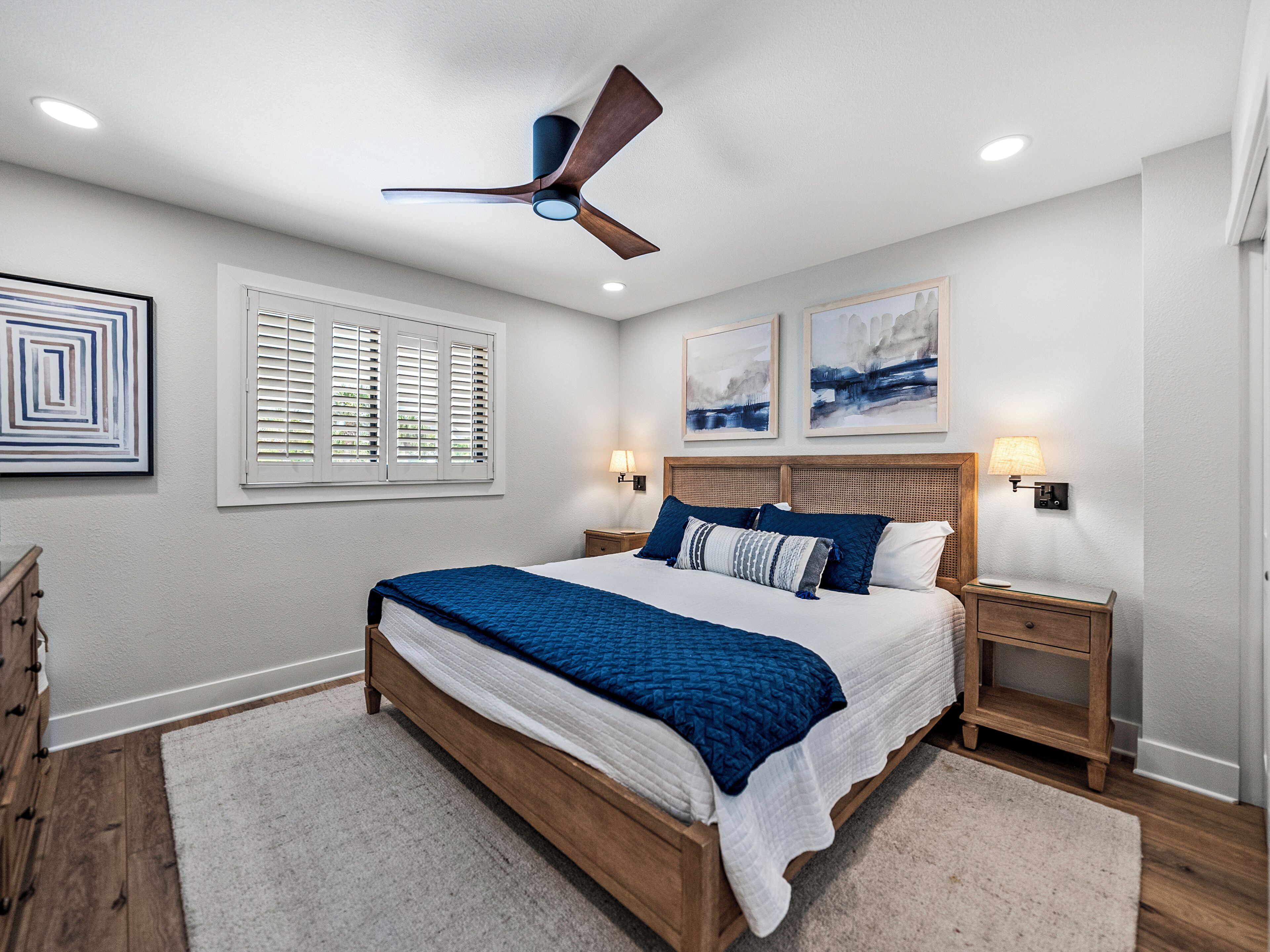 A bedroom with a large bed, wooden headboard, two bedside tables with lamps, a ceiling fan, and artwork on the walls.