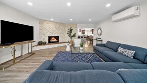 A modern living room with a flat-screen TV, a stone fireplace, a blue sofa, and a glass coffee table. - San Diego Lake home 4 Bd/Bath with heated pool, hot tub and golf! (Lakeside)