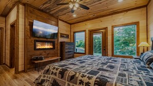 Une chambre à coucher dans un chalet en bois, dotée d’une cheminée, d’un téléviseur fixé au mur et de grandes fenêtres offrant une vue sur l’extérieur.