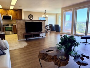A modern living room with a flat-screen TV, wooden flooring, a dining area with chairs, and a balcony with a view.