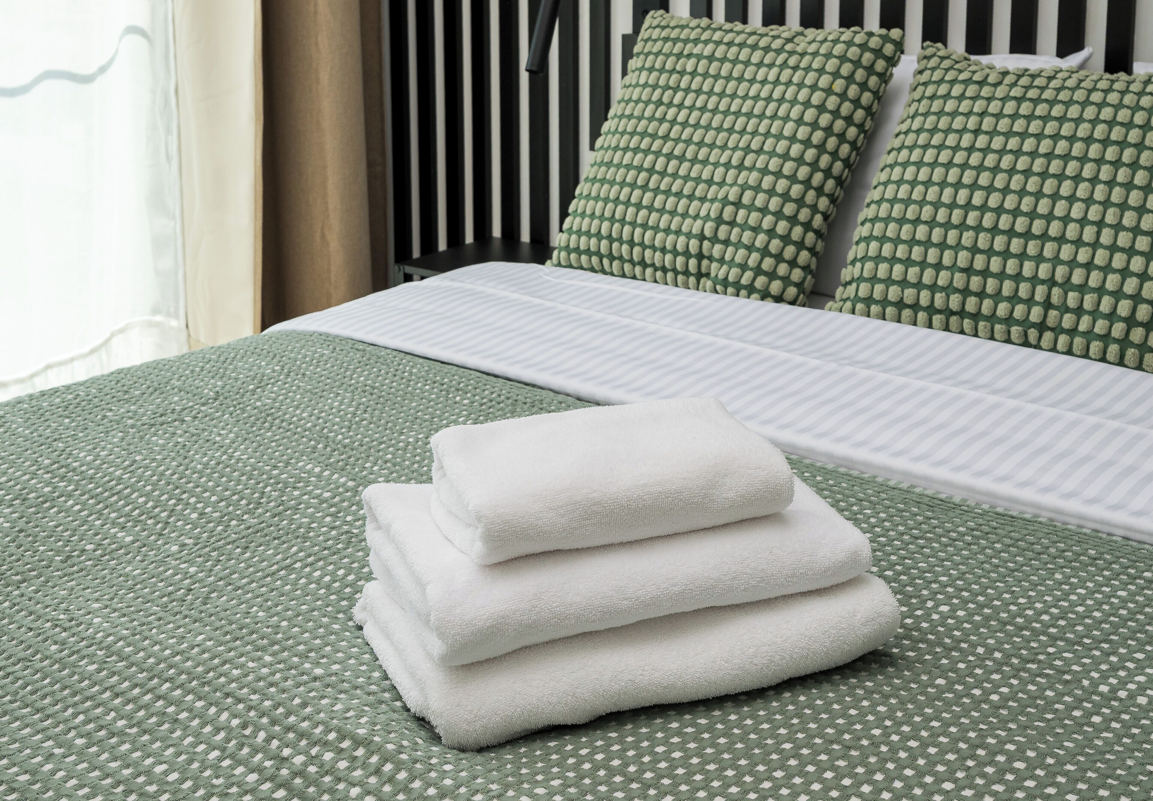 Una cama bien hecha con una colcha verde estampada y almohadas, y una pila de toallas blancas sobre la cama.