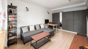 Un salon moderne avec un canapé d’angle gris, une table basse en bois, une étagère en métal noir agrémentée d’objets décoratifs, un petit bureau en bois et une armoire grise.