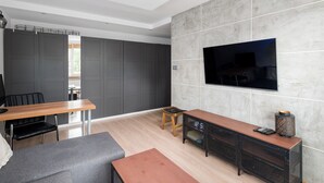 Un salon moderne avec un téléviseur fixé au mur, une table basse en bois, un canapé gris, un petit tabouret en bois et un meuble de rangement noir pour les appareils électroniques.