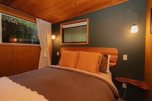 A bedroom with a wooden headboard, a window with curtains, a wall-mounted light, and a bedside table. - Mangamaunu Cottage, The Mangamaunu Cottage (Hapuku)