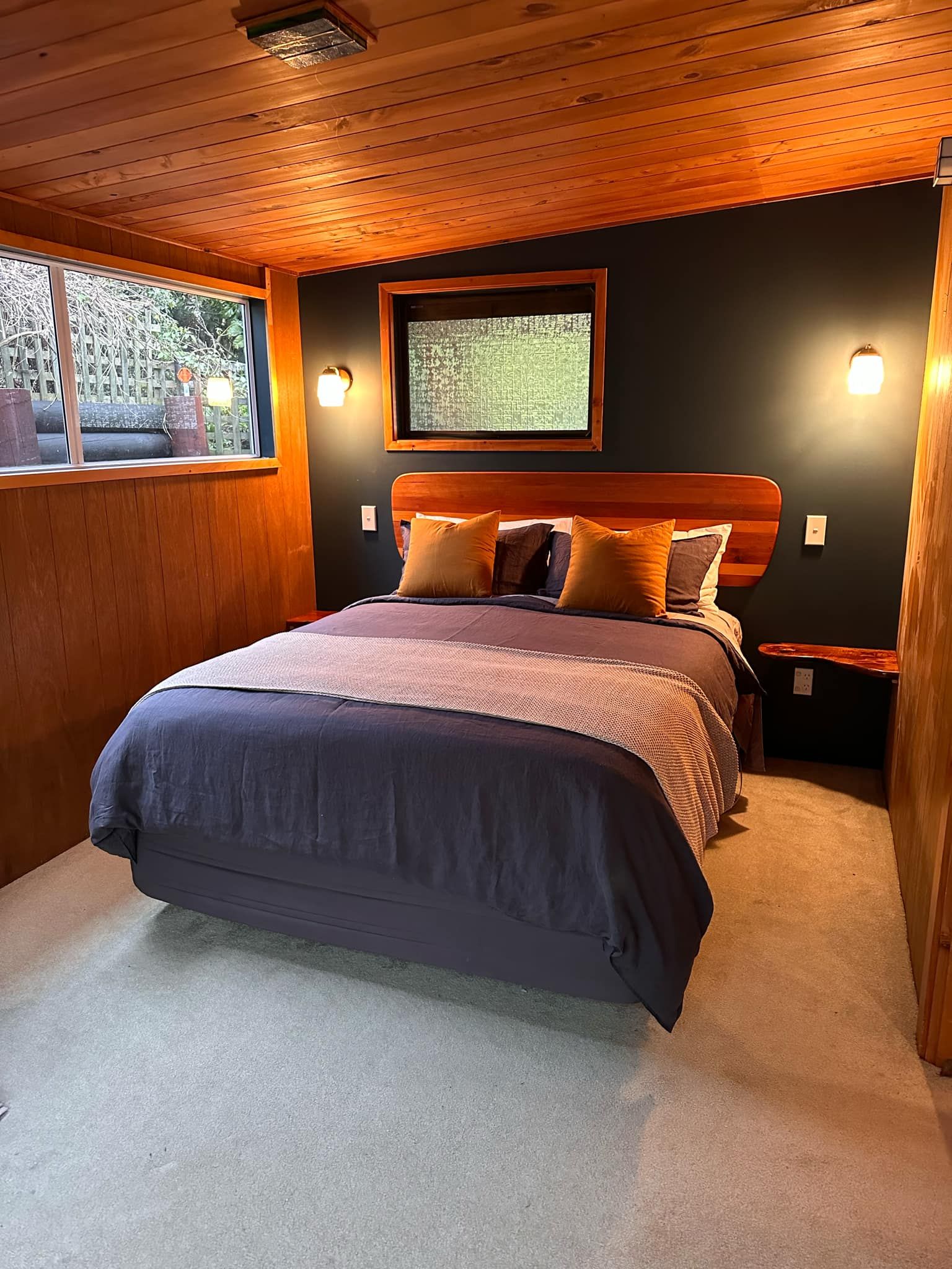 Un dormitorio con una cama grande, cabecera de madera, ventana con vistas y una lámpara fijada a la pared.