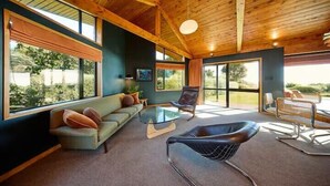 Een ruime woonkamer met houten plafond, grote ramen, een bank, een glazen salontafel en een moderne fauteuil.