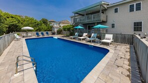 A rectangular swimming pool with a tiled deck, surrounded by lounge chairs and umbrellas, and a two-story building with a balcony in the background. - The Real Thing II - 6 bedroom semi-oceanfront with pool (Virginia Beach)