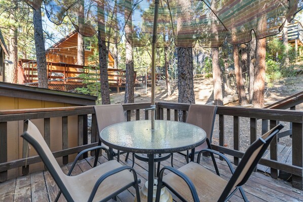 A wooden deck with a round glass table and chairs, surrounded by trees and a wooden cabin in the background. - Mountain Bliss (Big Bear City)