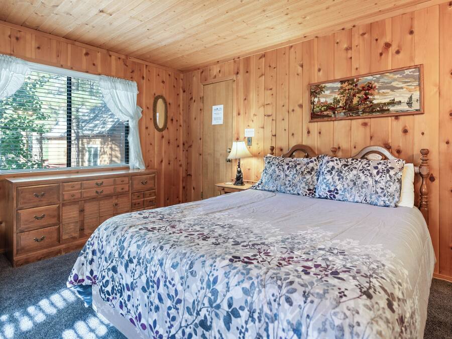 A wooden-paneled bedroom with a bed, dresser, window with curtains, and a painting on the wall.