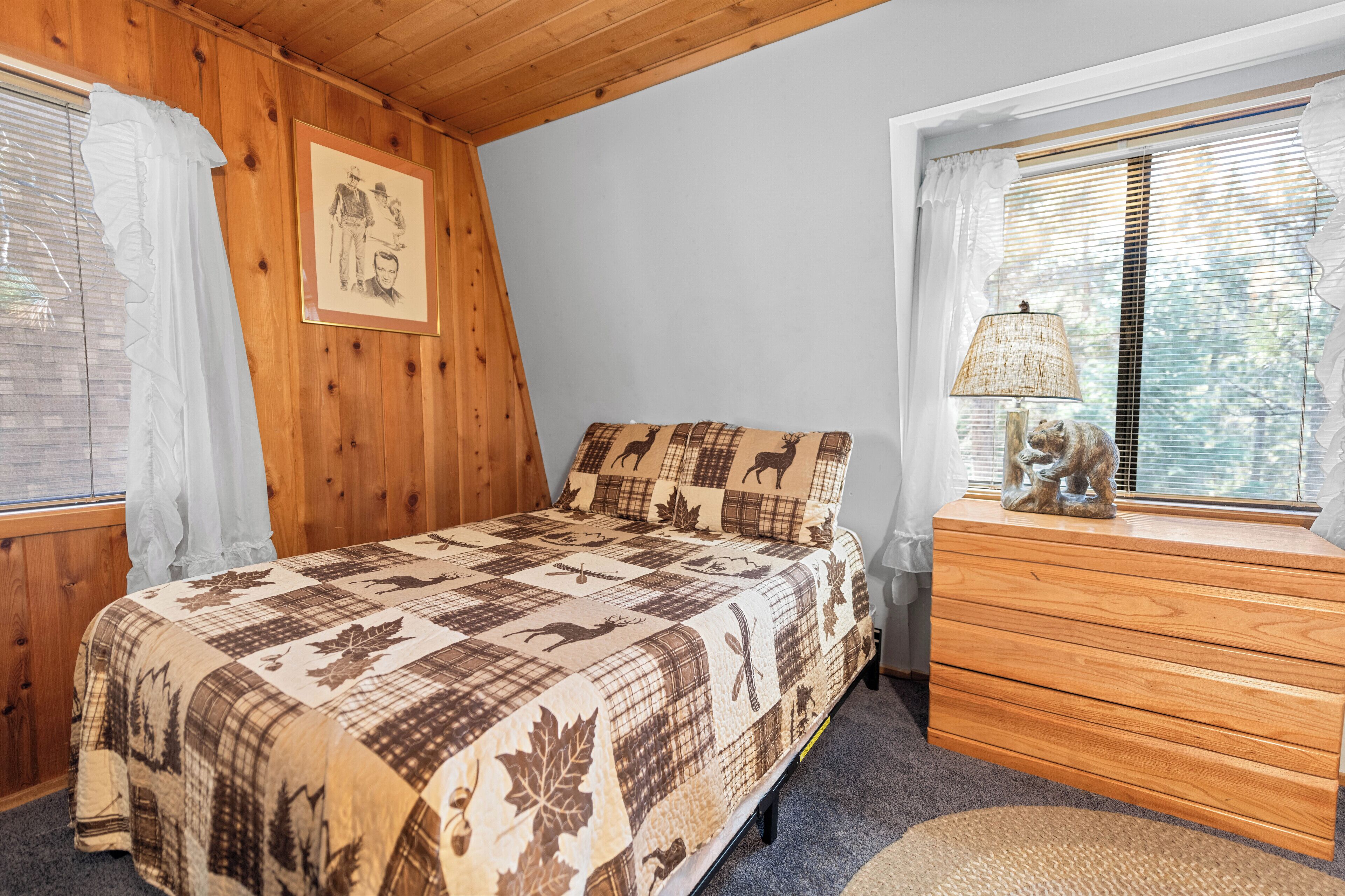 Un dormitorio con techo de madera, una cama con colcha estampada con motivos animales, un aparador con lámpara y una ventana con persianas.