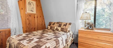 Un dormitorio con techo de madera, una cama con colcha estampada con motivos animales, un aparador con lámpara y una ventana con persianas.