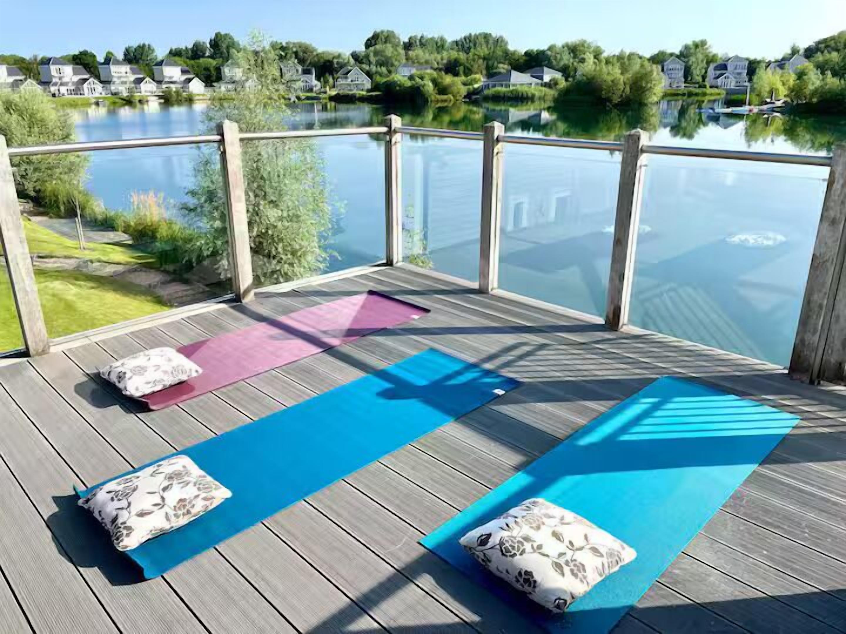 Une terrasse dotée de tapis de yoga et de coussins, offrant une vue sur un lac paisible, avec des maisons en arrière-plan.