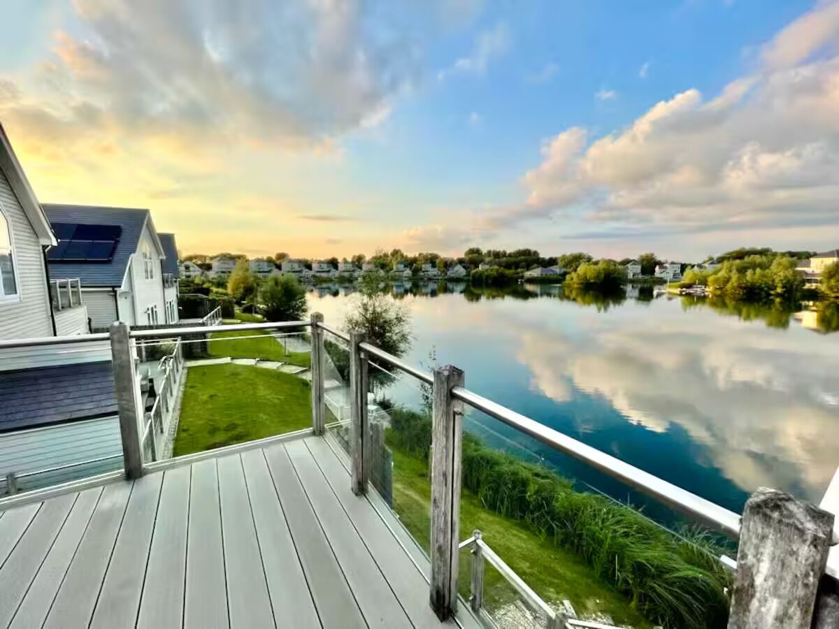 A wooden deck with a railing overlooking a calm lake, surrounded by greenery and residential buildings.