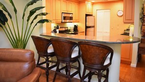A kitchen with wooden cabinets, a stainless steel refrigerator, and a microwave. There are three bar stools with cushioned seats. A large green plant is visible in the corner. - Wild Oak Bay - 3/2 Beaches 15 minute drive (Bradenton)